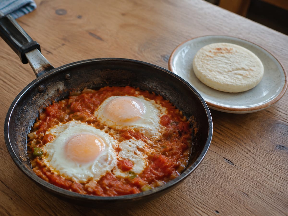 Huevos cocidos sobre hogao de tomate y cebolla acompañados con arepa